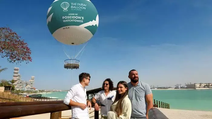 Four people stand near a boardwalk by the water, looking toward a large tethered gas balloon hovering in the sky at The Dubai Balloon at Atlantis.