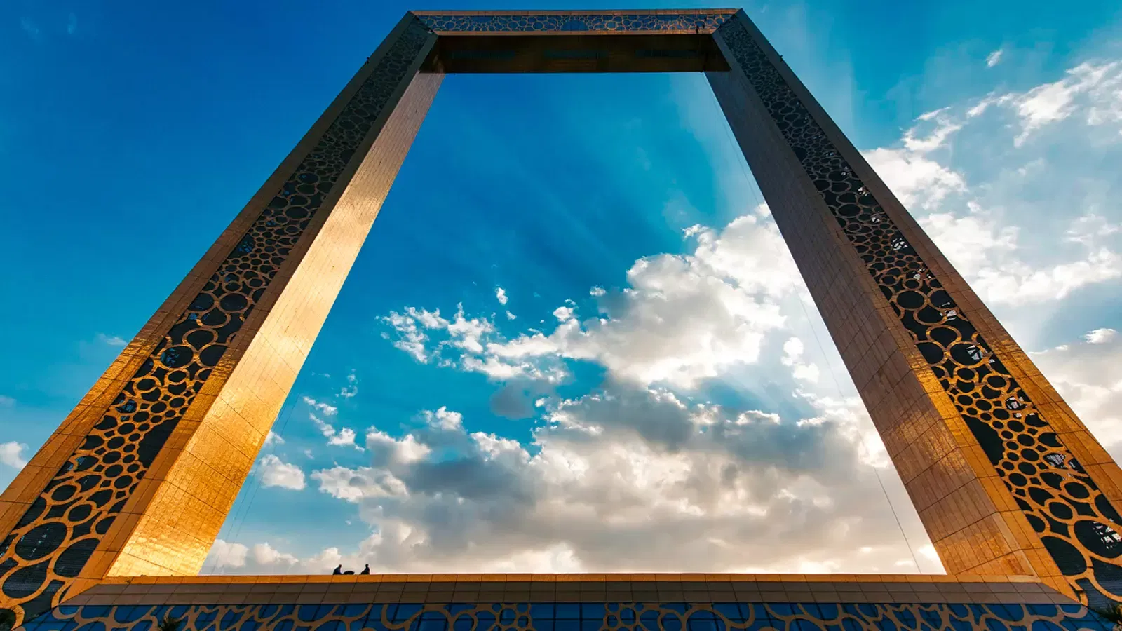 A low-angle daytime view between the two golden columns, with two small figures standing on the lower crossbar, at Dubai Frame
