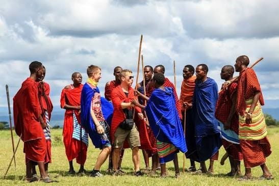 A group of tourists in traditional shúkà fabrics dancing outdoors in a circle with Maasai hosts.