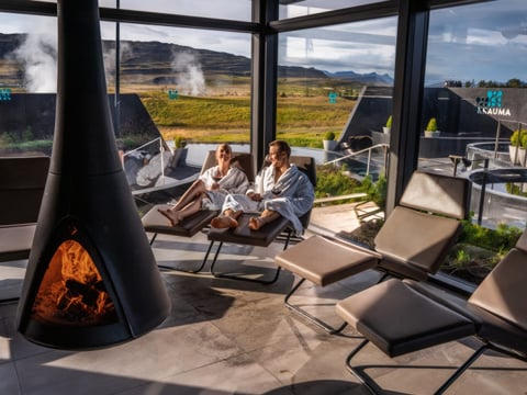 Two guests in white robes reclining on loungers inside Krauma's glass-walled relaxation room, with a suspended wood-burning fireplace in the