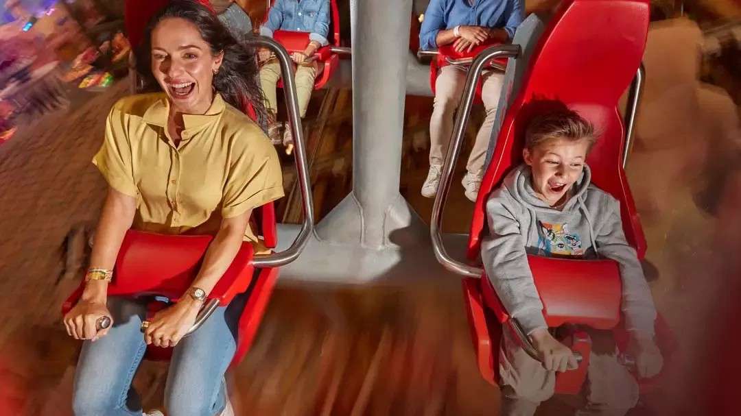A woman and a boy ride in red, forward-facing seats on an indoor amusement park ride at Warner Bros. World Abu Dhabi Theme Park.