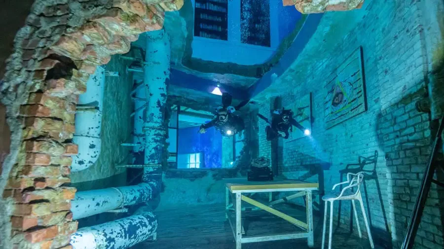 Two scuba divers explore an underwater room featuring brick walls, pipes, and furniture at Deep Dive Dubai.