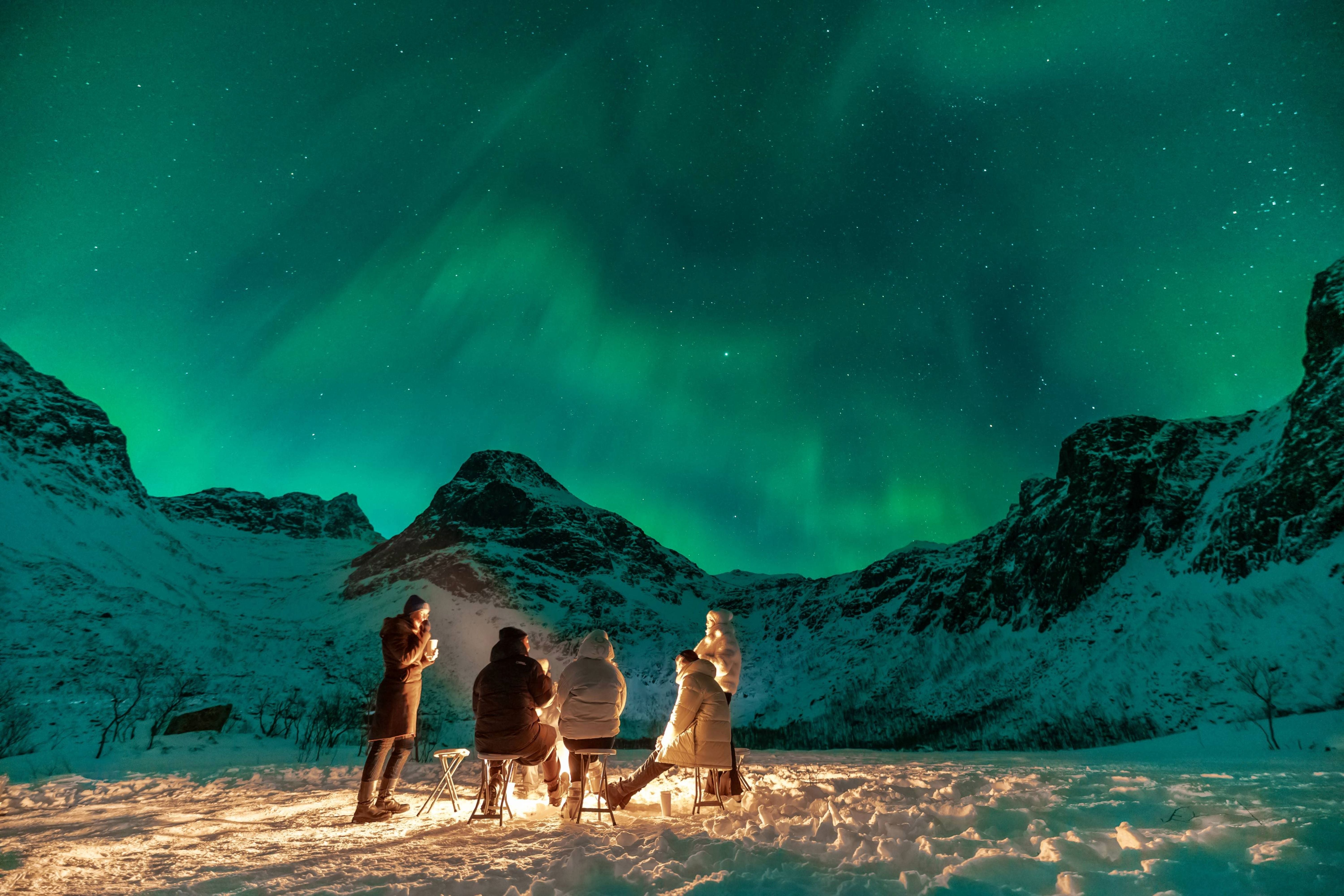 Small group of travelers sitting around a warm campfire in a snow-covered mountain valley beneath the green Aurora Borealis.