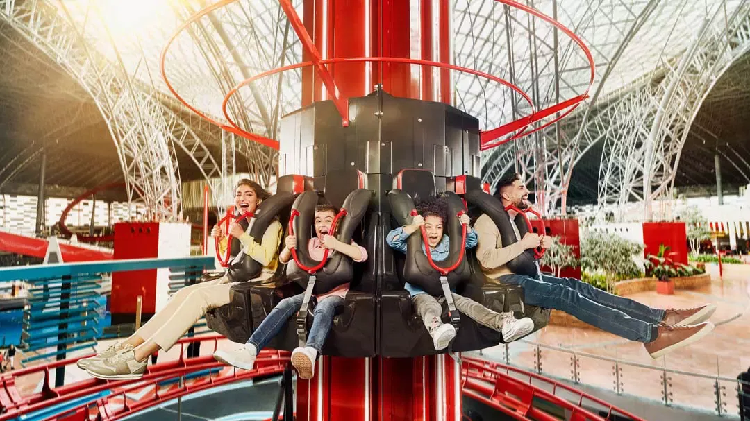 Four riders with legs dangling from a black drop-tower seat block, the white lattice roof of the park arching above them, at Ferrari World Abu Dhabi