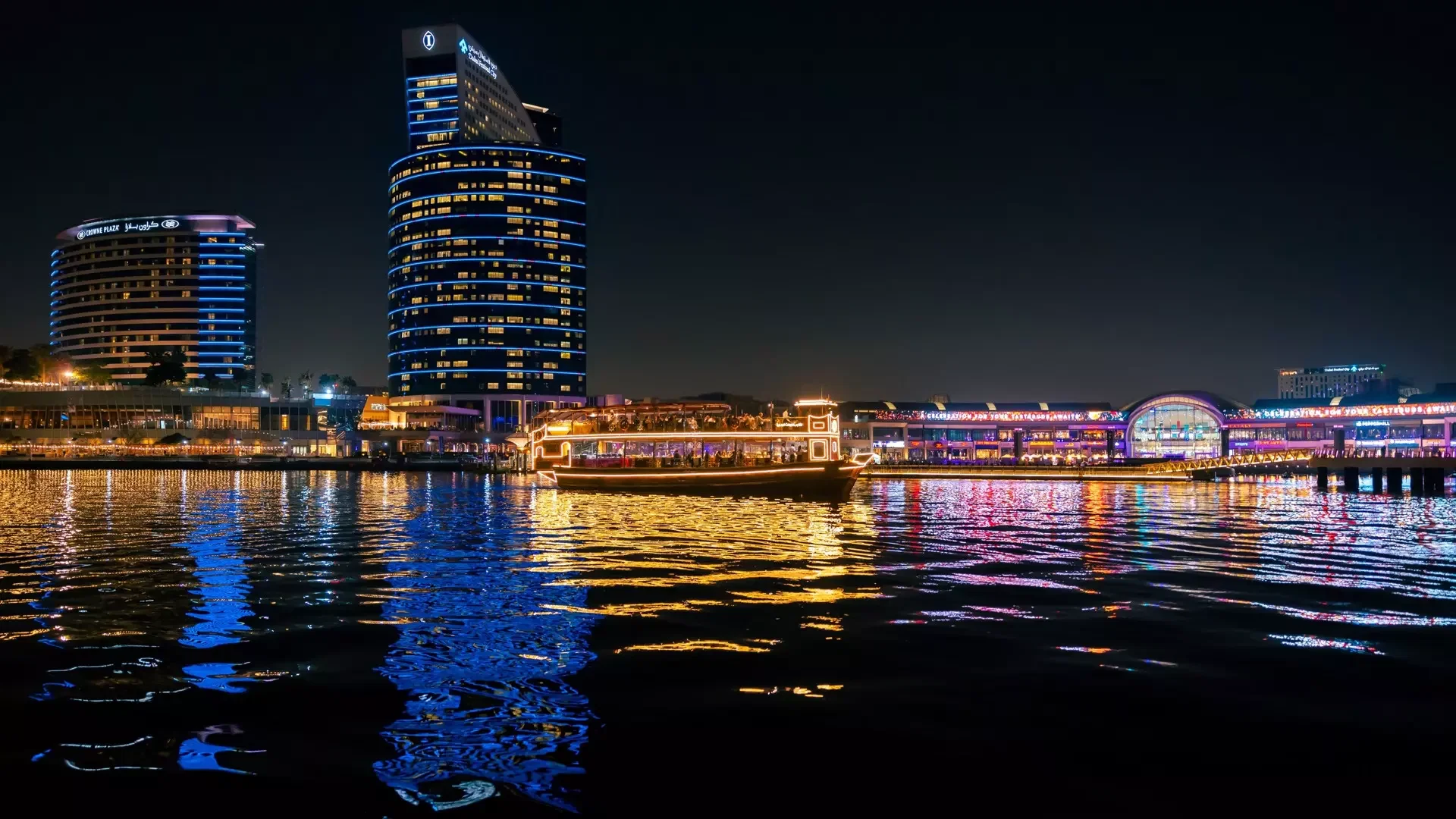 A lit-up traditional boat cruises on a dark river at night, with brightly illuminated modern high-rise buildings and waterfront structures reflecting in the rippling water at Dubai Water Canal Dhow Cruise.