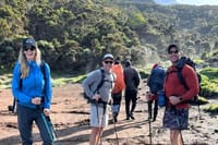 Three smiling hikers standing on a sunny mountain trail with backpacks and trekking poles.