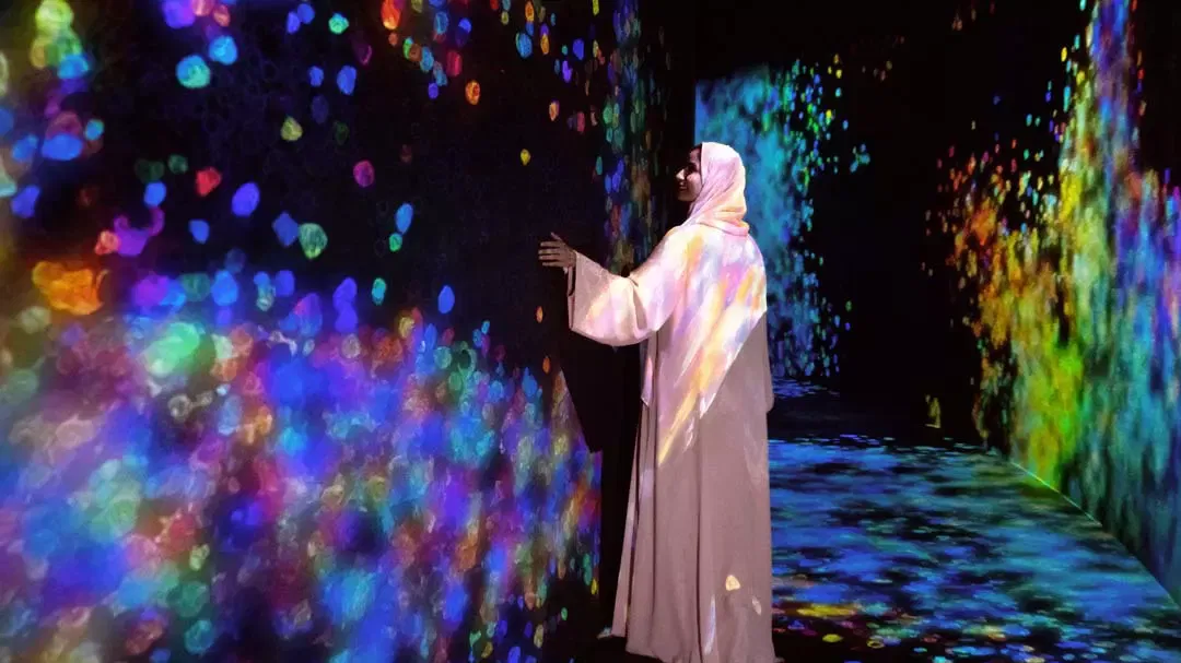 A woman in a hijab gently touches a dark wall covered in colorful floating light circles at TeamLab Phenomena.