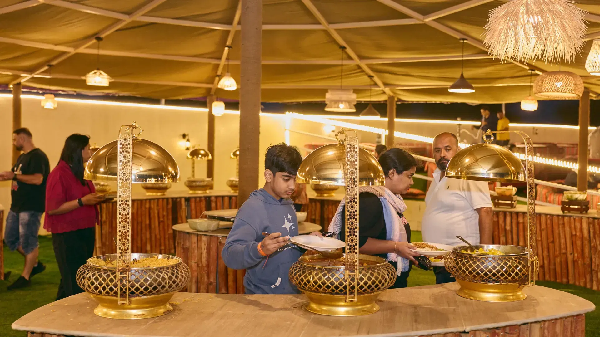 People gather around a buffet line with golden chafing dishes under a lit tent at Desert Safari + Al Faqaa Camp.