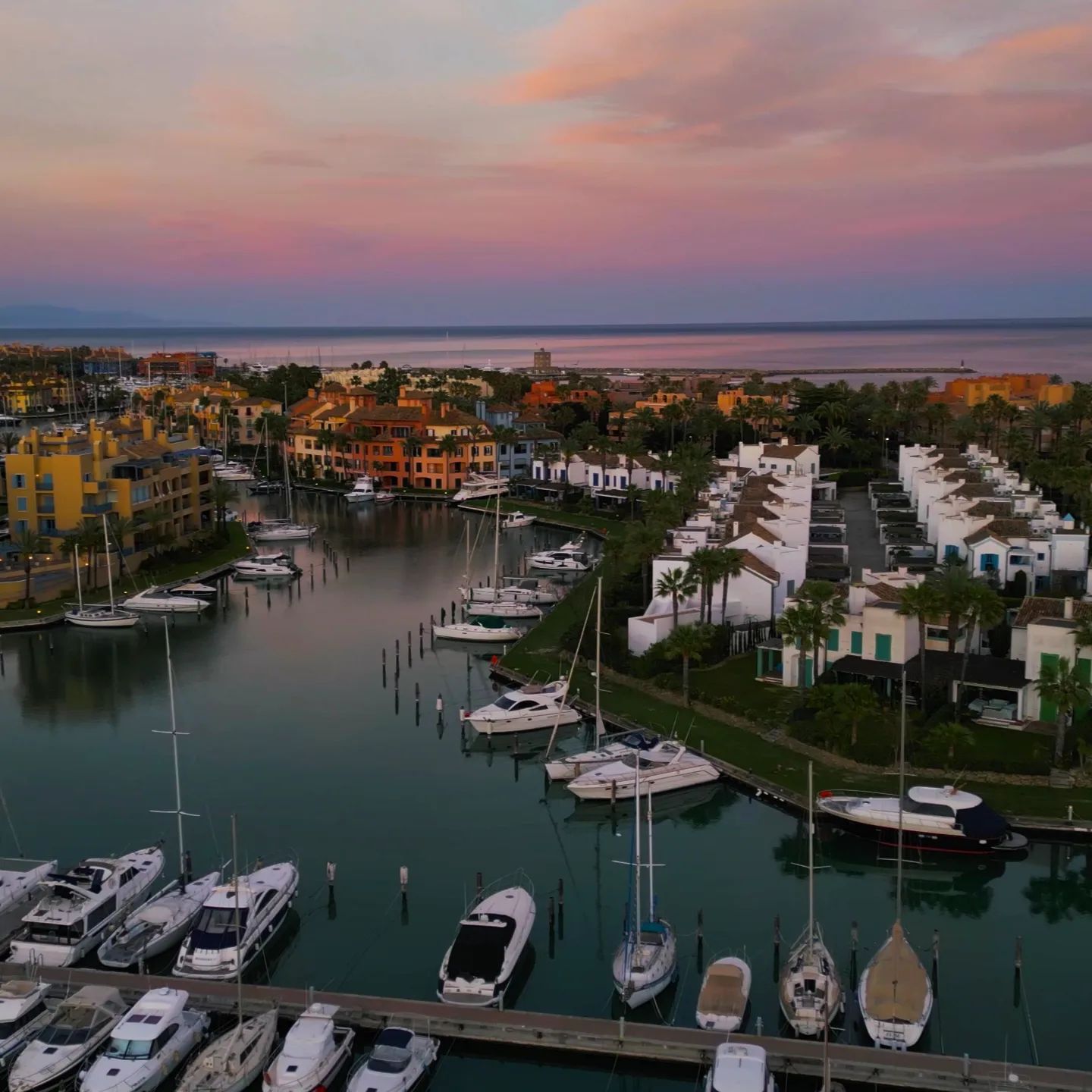 Vista aérea del puerto de Sotogrande al atardecer, con el sol brillando entre las nubes antes de ocultarse en el horizonte, reflejando su lu