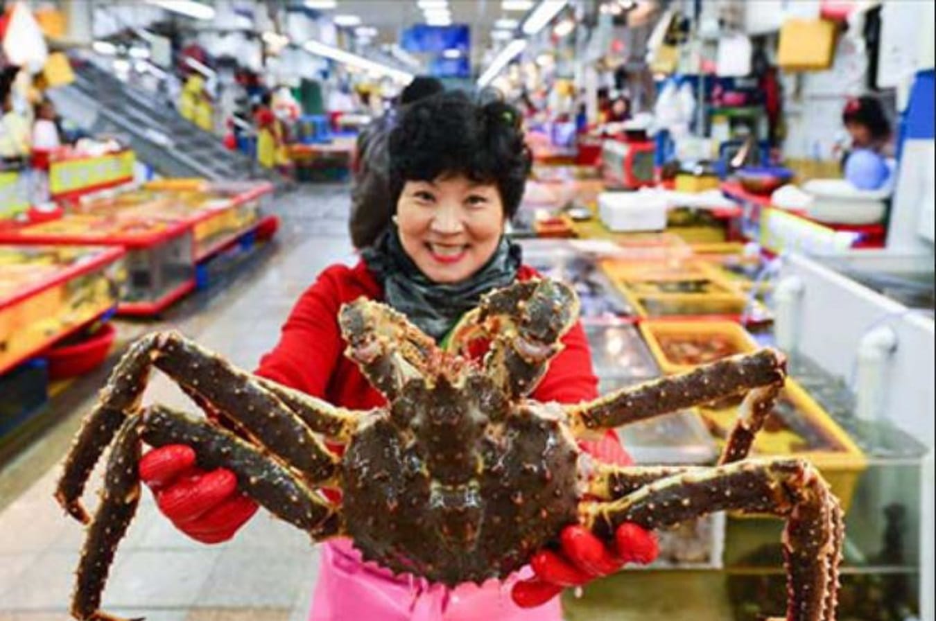 A vendor holds up a large king crab at Jagalchi Fish Market, with stalls of fresh seafood in the background.