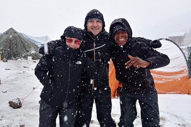 Three people in black winter jackets smiling and hugging during a snowfall at a mountain camp with tents in the background.