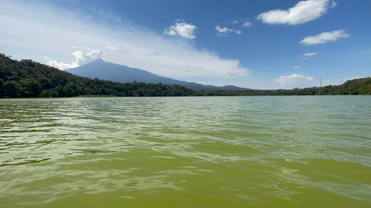 Panoramic landscape view of Lake Duluti's green water with Mount Meru under a blue cloudy sky.