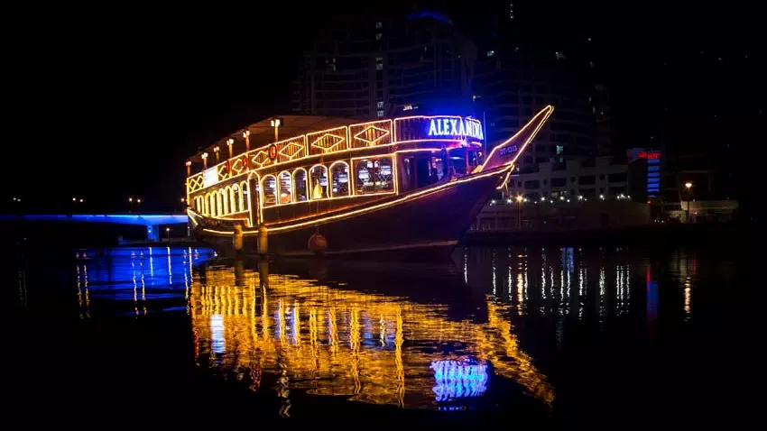 A large wooden boat illuminated with bright golden lights is docked on dark water at night, with its warm reflection shimmering on the surface at Alexandra Dhow Cruise in Dubai Marina.
