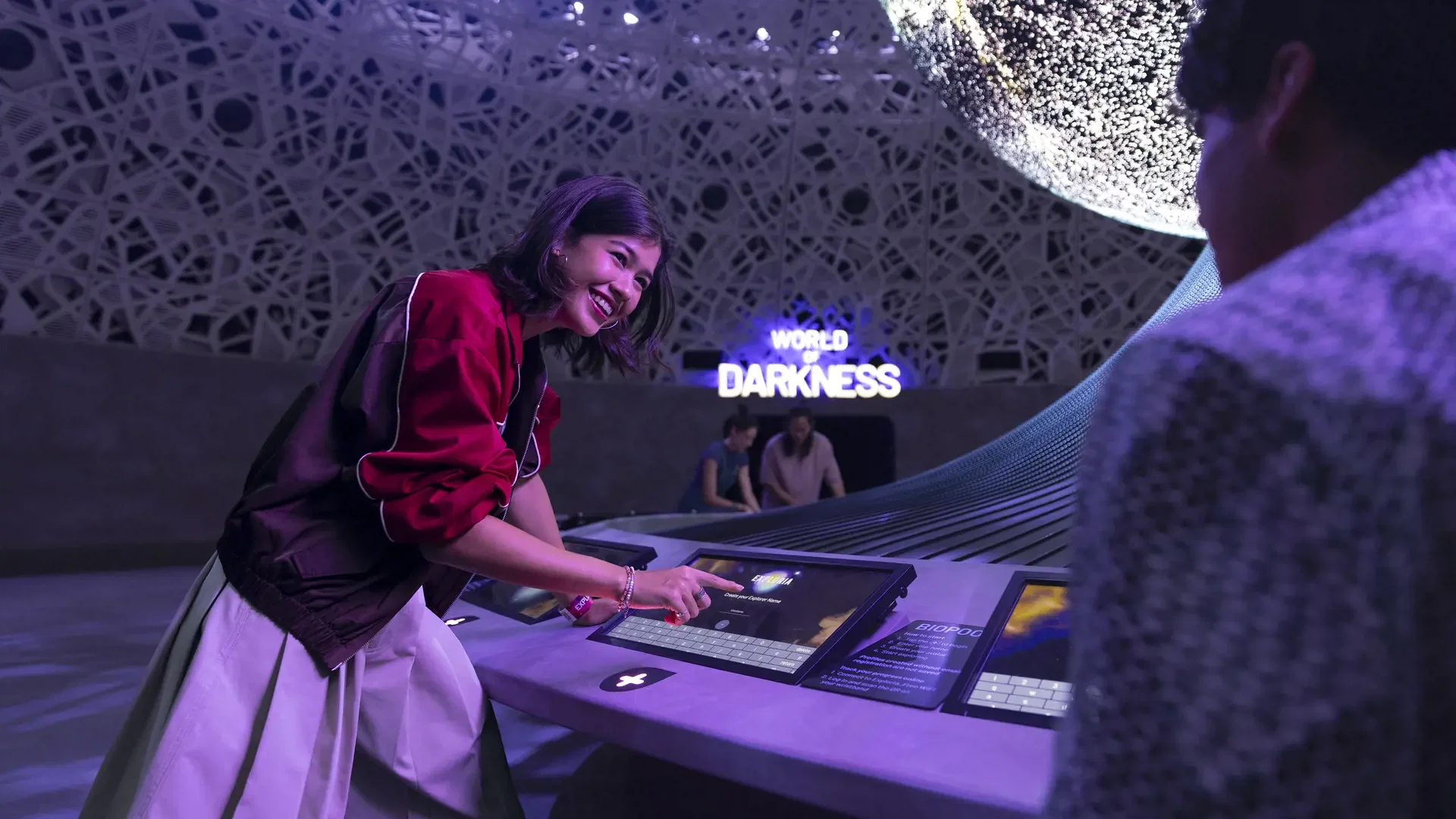A smiling woman in a red jacket types on an interactive digital console beneath a lit sign reading
