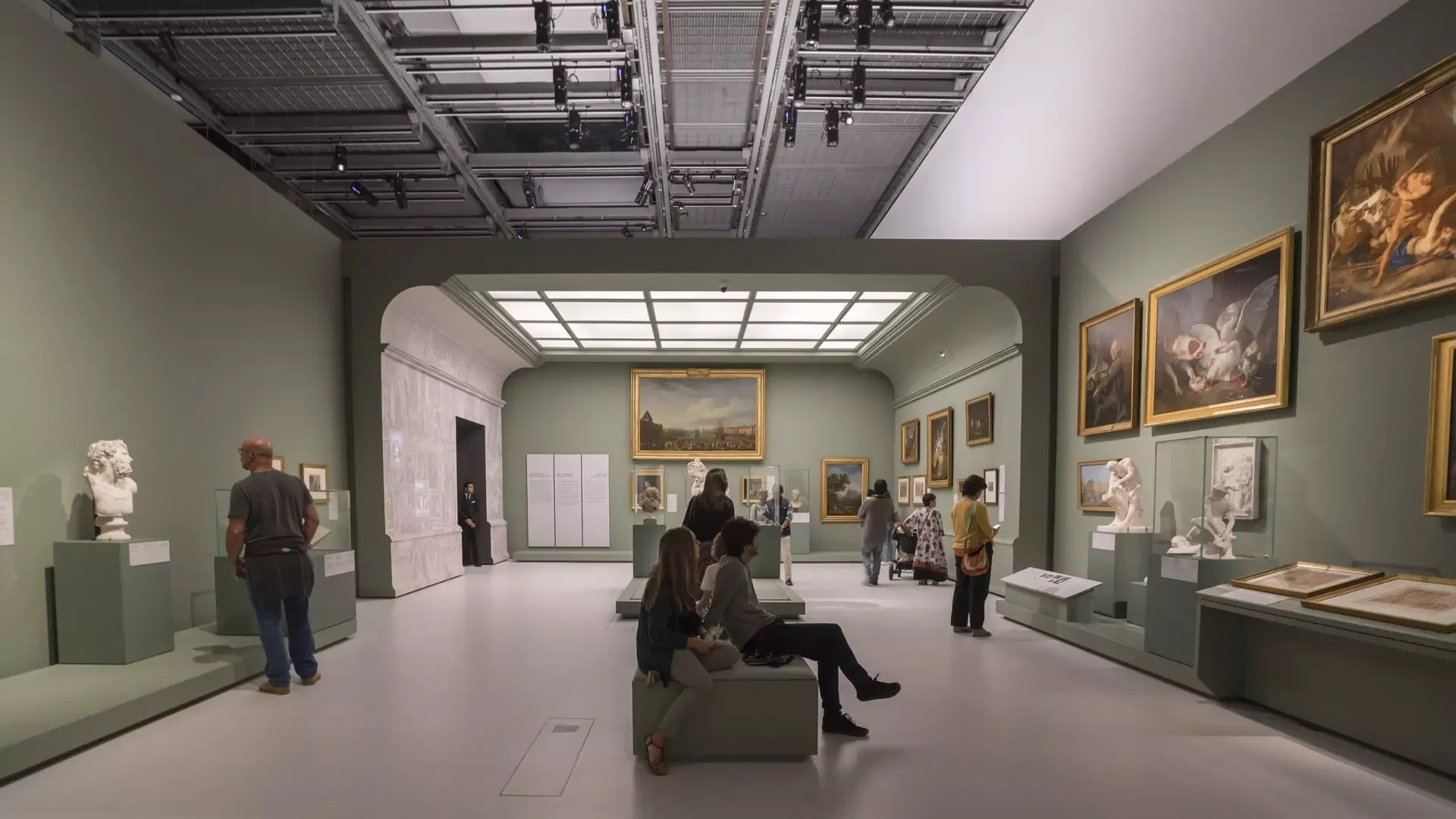 Visitors seated and standing in a sage-green gallery hung with gilt-framed paintings and lined with marble busts at Louvre Abu Dhabi