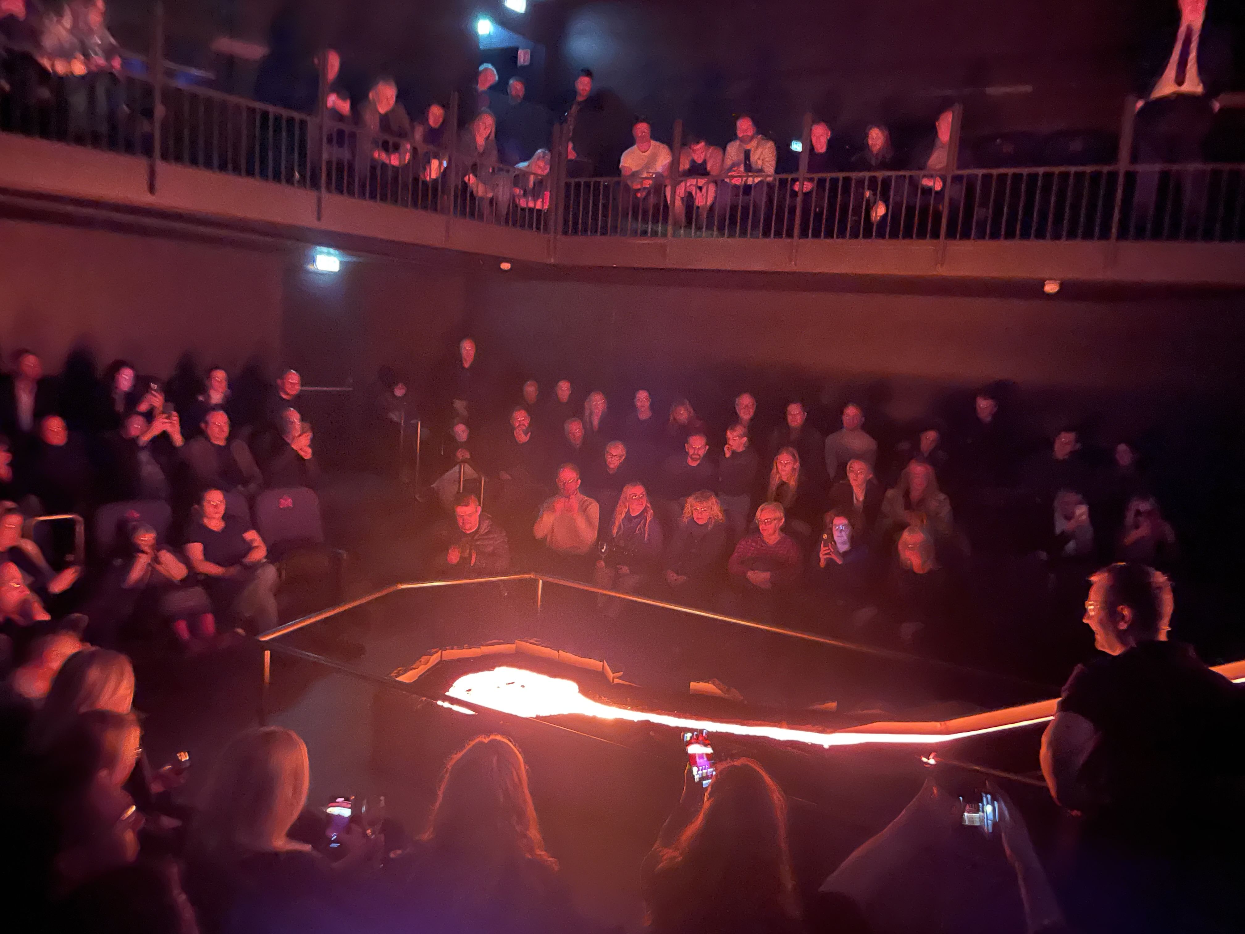 Smiling audience members sit close to a steaming lava pit, lit in red and white as molten rock cools and releases steam.