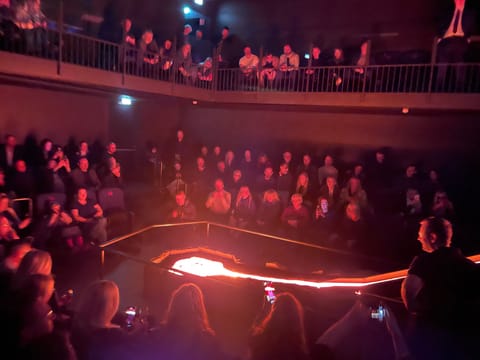 Smiling audience members sit close to a steaming lava pit, lit in red and white as molten rock cools and releases steam.