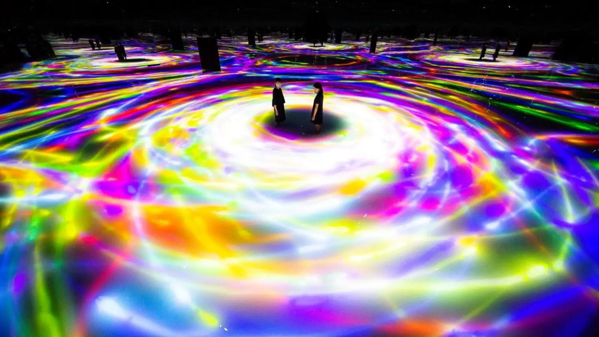 Two visitors stand in a shallow pool of water reflecting swirling, vibrant projections of multicolored light trails at COMBO: Tokyo Skytree + teamLab Planets TOKYO Tickets.
