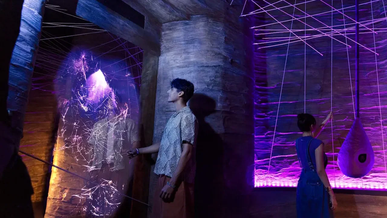 A young man and woman explore a rocky cavern with purple lighting and glowing string art installations at Exploria in Mandai Singapore Ticket.