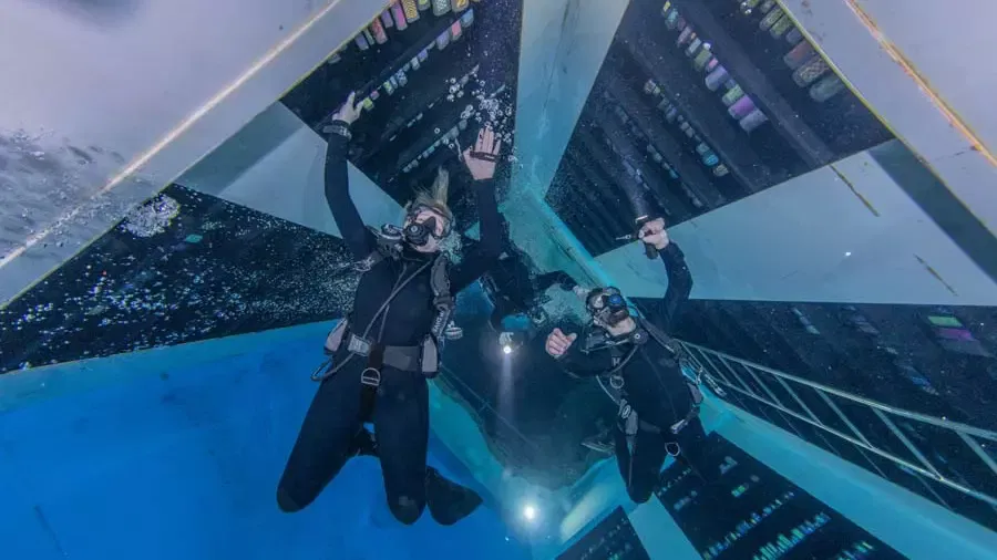 Three scuba divers in black wetsuits explore the bright blue depths, surrounded by geometric underwater architecture and bubbles at Deep Dive Dubai.