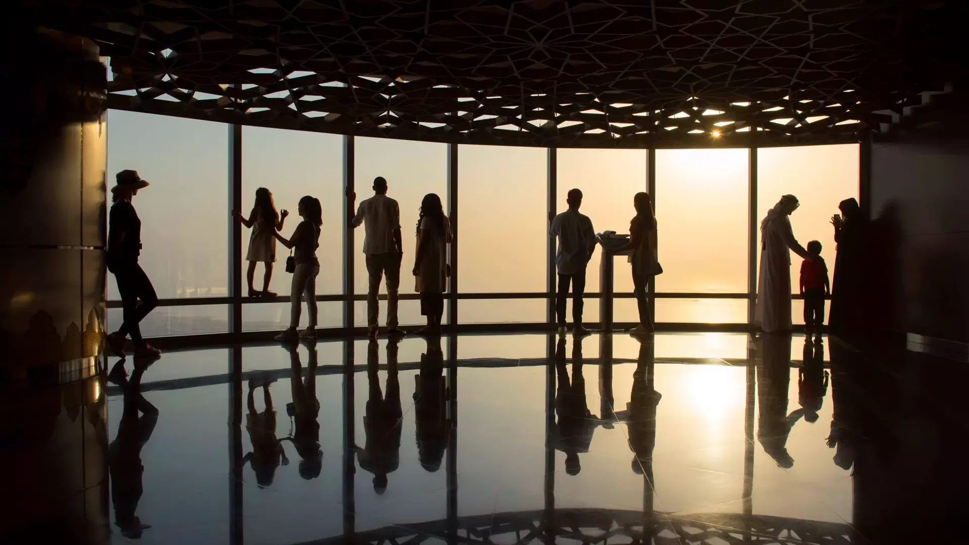 Visitors silhouetted against a hazy orange sunset along the curved window wall of the observation deck, their reflections in the polished floor beneath a patterned ceiling, at Burj Khalifa