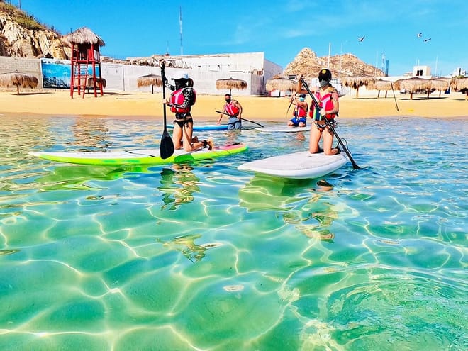 People learning paddle boarding in shallow clear water at a beach in Cabo San Lucas wearing life jackets