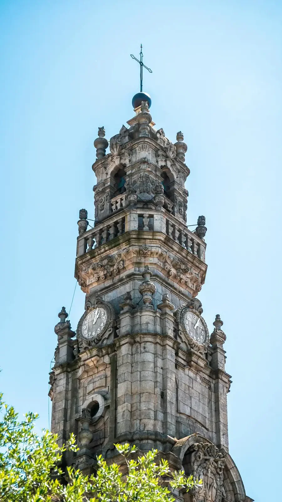 A close-up captures the upper section of a carved stone tower featuring clock faces and a cross on top against a clear blue sky at Clérigos Tower & Museum Ticket.