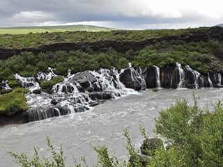Ground-level view of Hraunfossar waterfalls under overcast skies, with multiple cascades spilling over black lava rock into a grey-blue rive