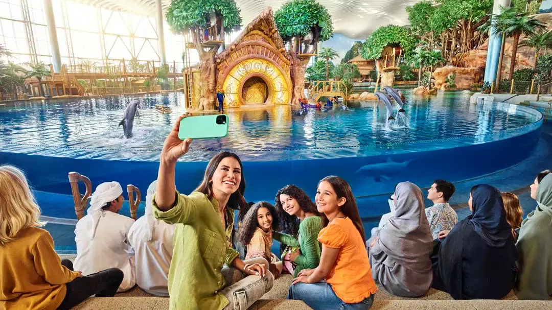 A smiling woman holds up a phone to take a selfie with a group of diverse visitors watching dolphins jumping in a large pool at Sea World Abu Dhabi.