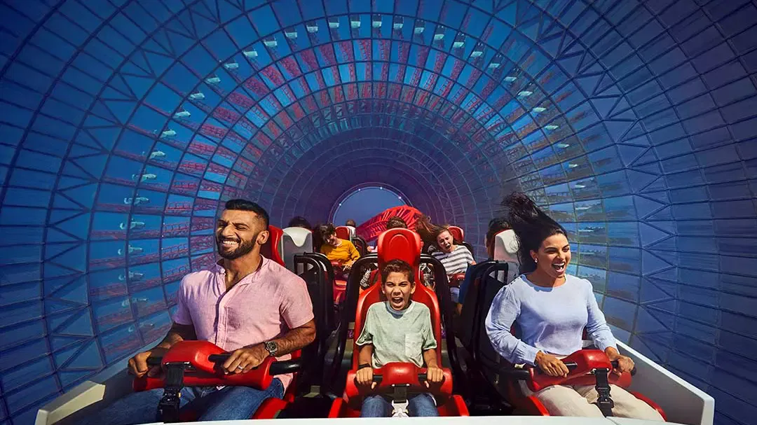A family screaming with hands on the harnesses as their coaster car races through a blue tunnel lined with red steelwork at Ferrari World Abu Dhabi