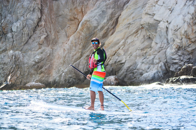 Man stand up paddle boarding in Cabo San Lucas near rocky cliffs wearing life jacket and sunglasses