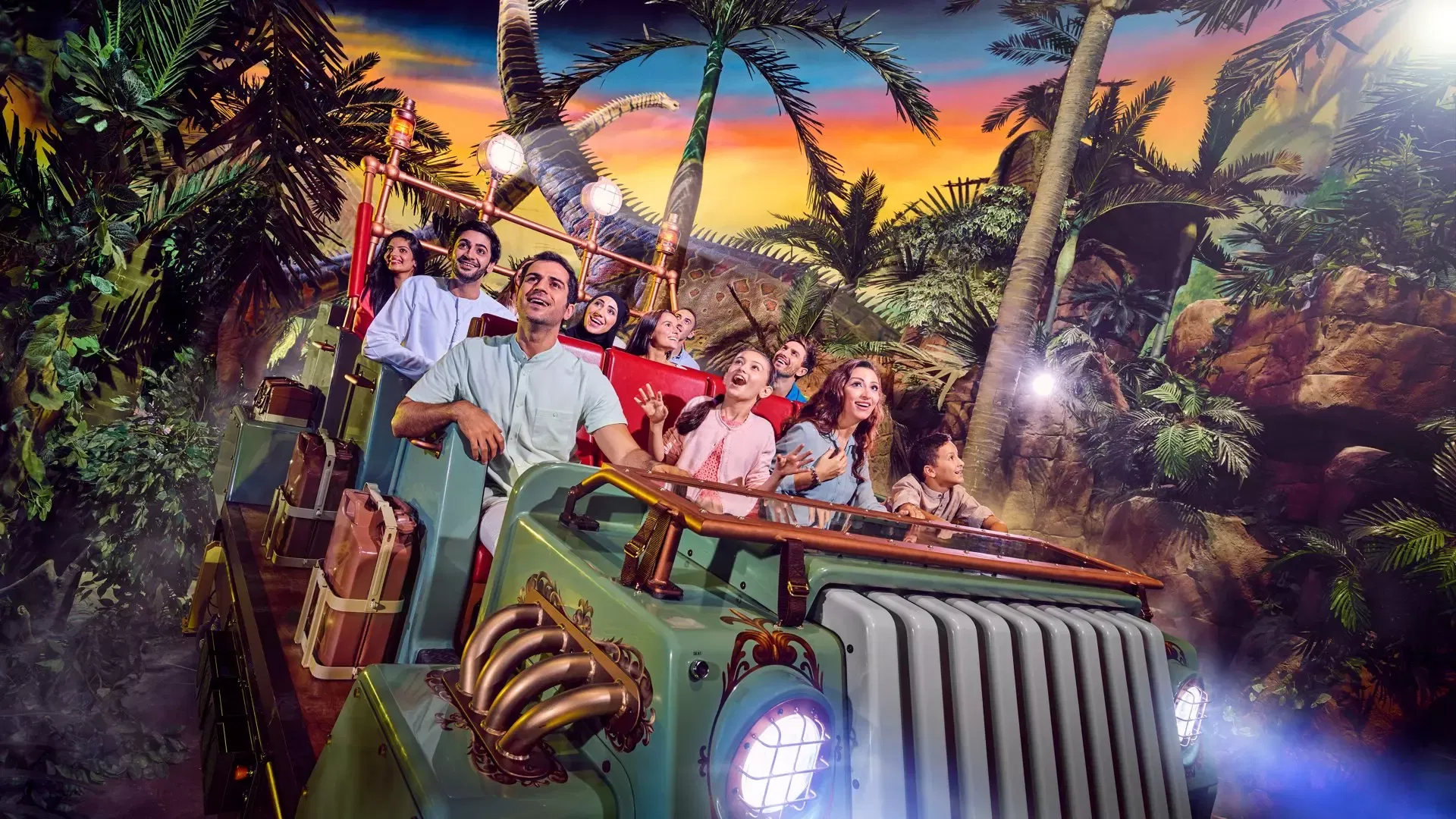 A group of smiling passengers on a green safari vehicle ride through a jungle setting with large dinosaur models at IMG Worlds of Adventure Theme Park.