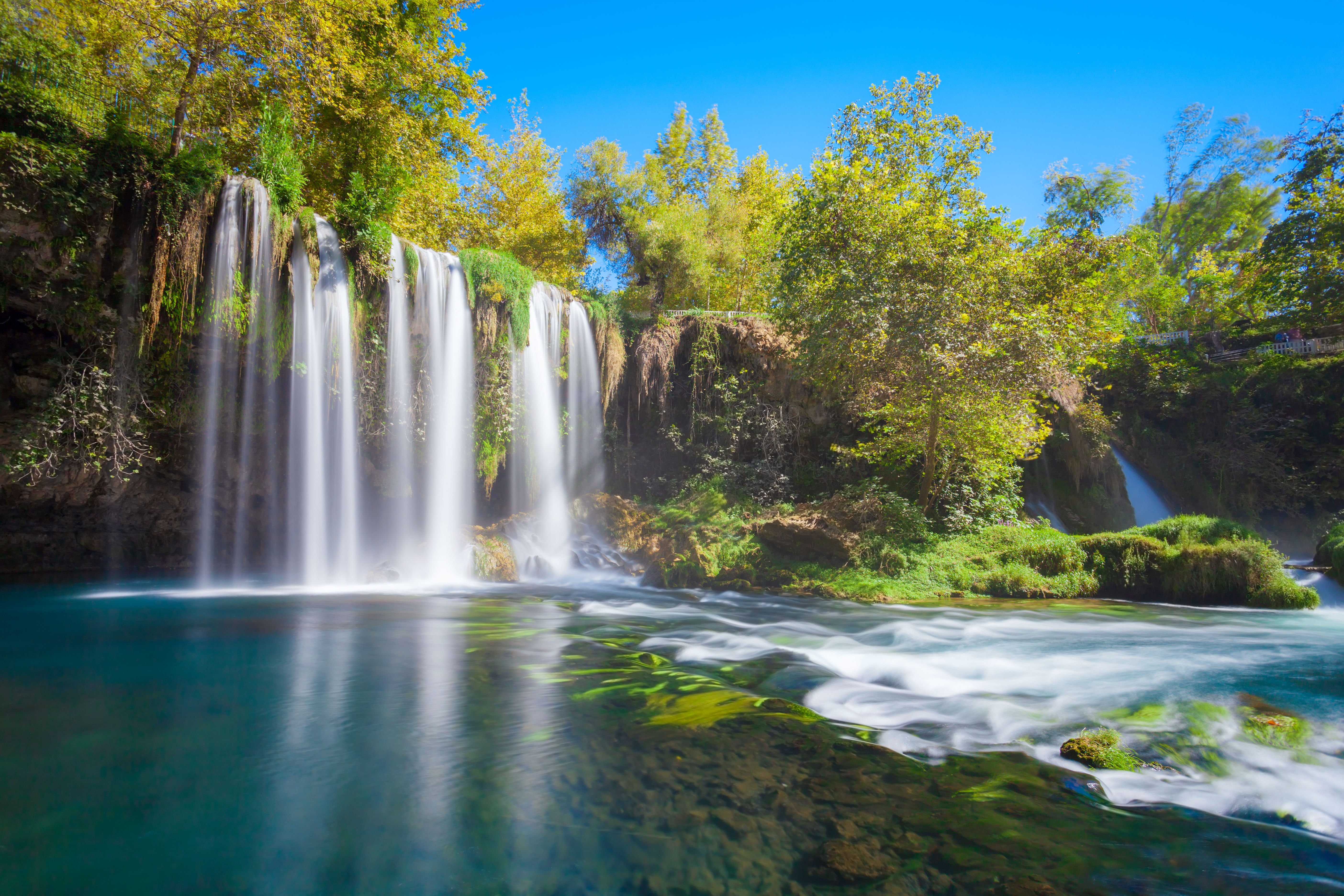 “View of Duden Waterfall in Antalya flowing over rocky cliffs into a green park area with trees and walking paths.”