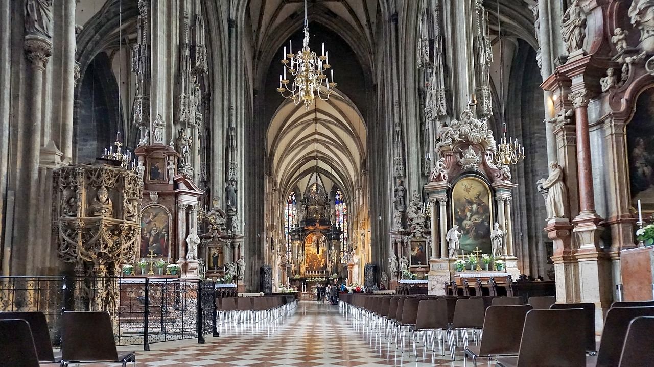 View down the nave of St. Stephen’s Cathedral showing tall Gothic arches, chandeliers, ornate altars, and rows of chairs.