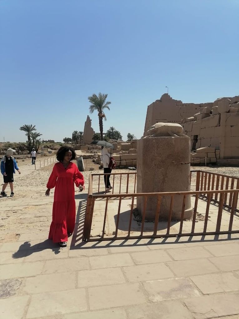 Une femme vêtue d'une robe rouge se tient près d'un ancien pilier de pierre clôturé parmi les ruines du temple de Karnak, avec d'autres touristes et des palmiers dans