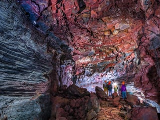 Small group of visitors in helmets standing inside the illuminated Vidgelmir lava tube, dwarfed by towering walls of layered grey and deep r