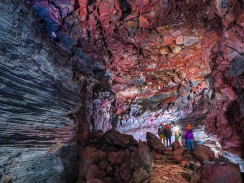 Small group of visitors in helmets standing inside the illuminated Vidgelmir lava tube, dwarfed by towering walls of layered grey and deep r