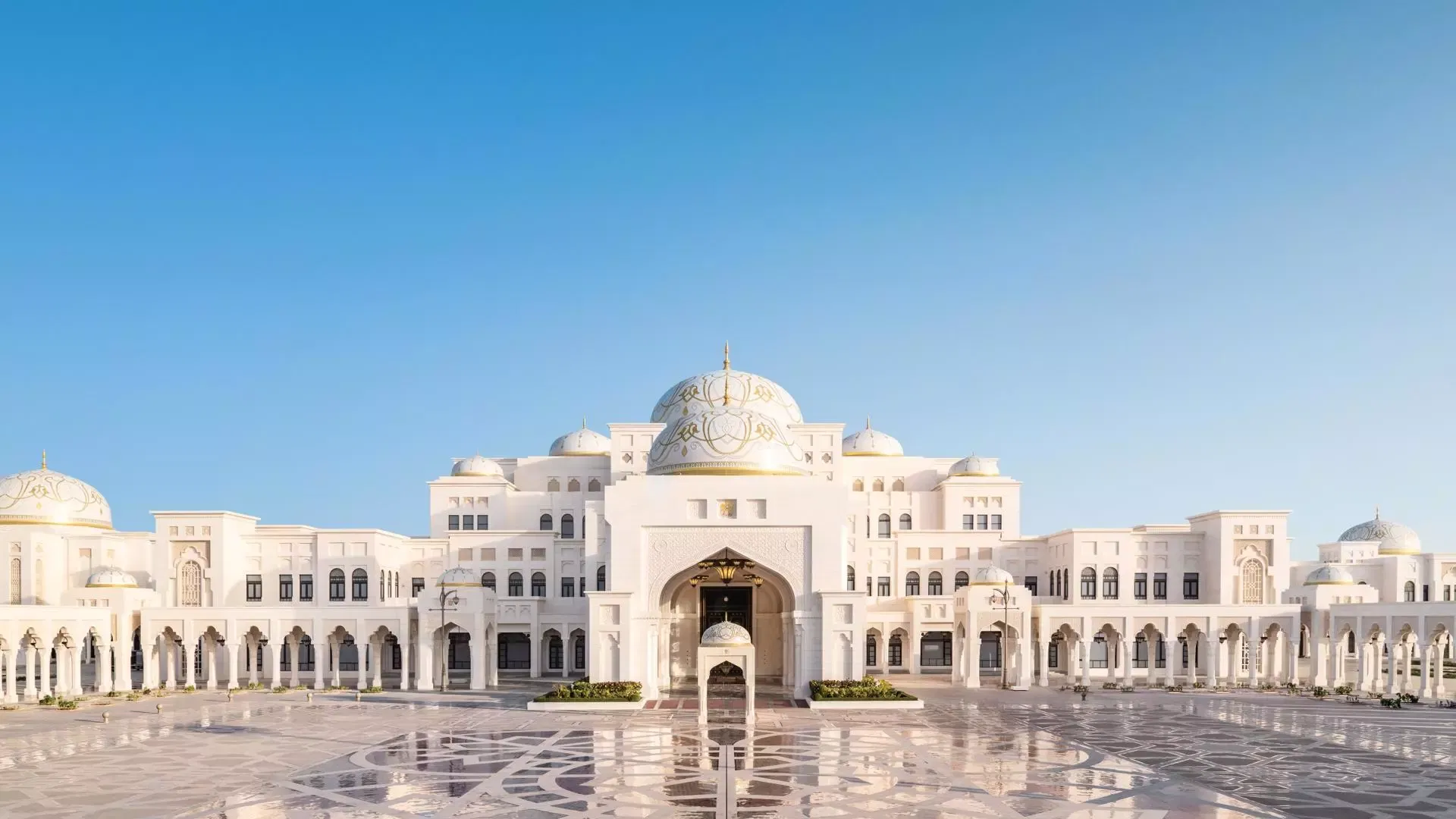 The sprawling white stone exterior features multiple domes, arched porticoes, and a vast paved courtyard under a clear blue sky at Qasr Al Watan, Royal Palace.