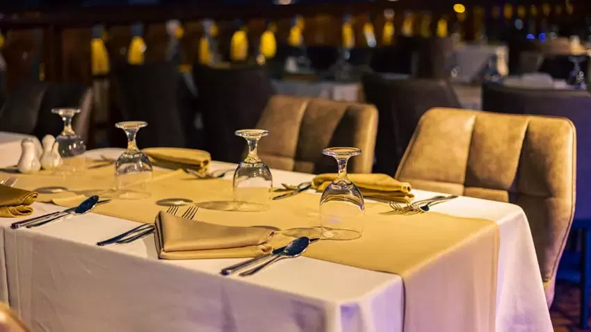 A close-up view of a dining table covered in a white tablecloth and gold runner, featuring neatly folded napkins, cutlery, and water glasses at Alexandra Dhow Cruise in Dubai Marina.