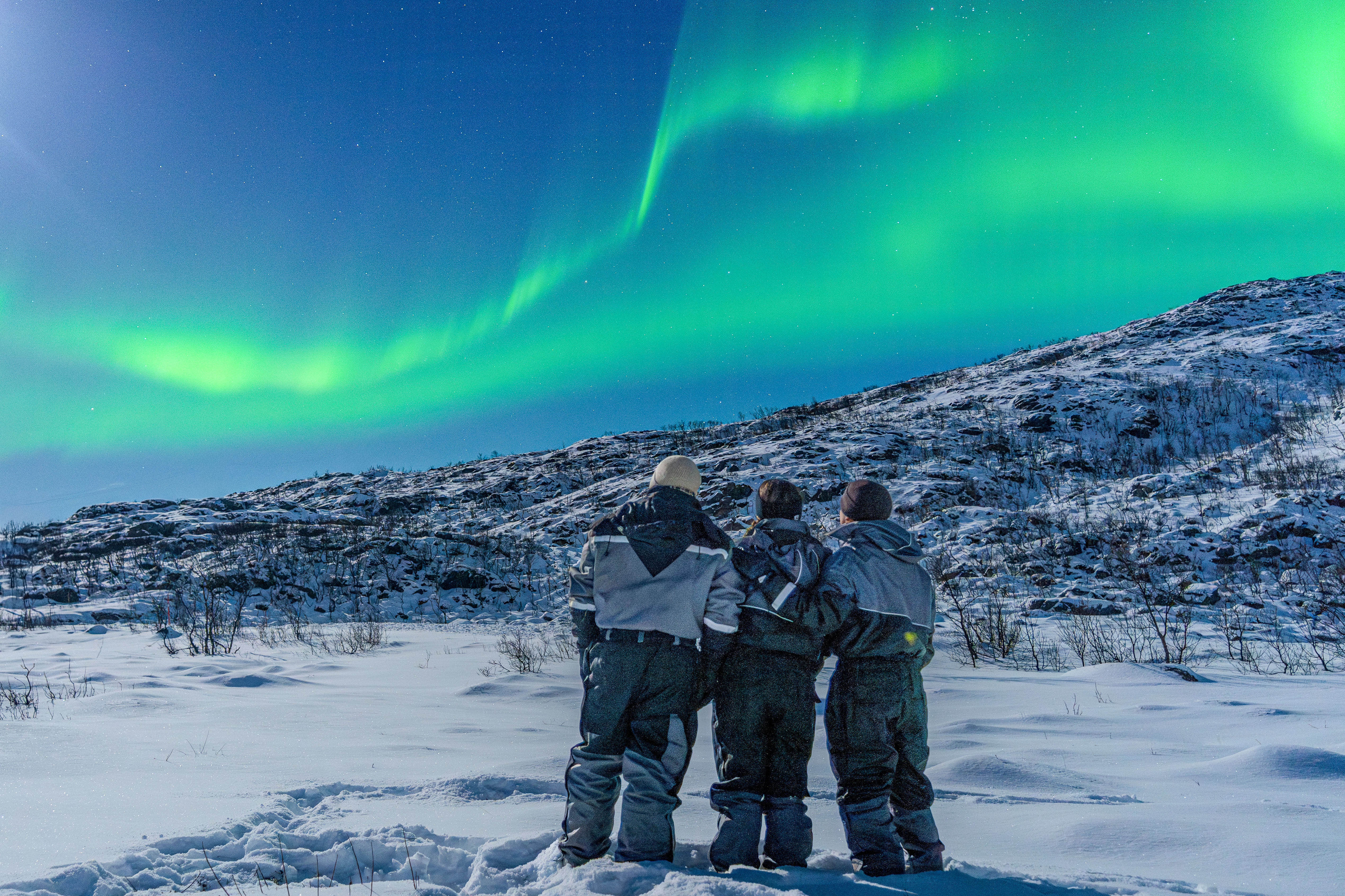 Three people standing in snowy Arctic landscape near Tromsø, watching bright green Northern Lights during a guided tour in Northern Norway.
