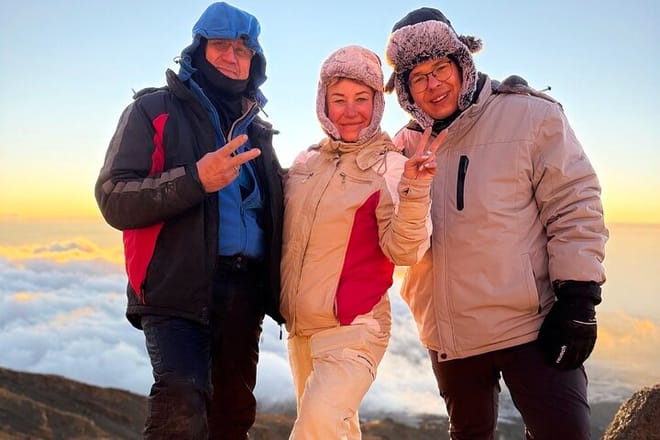 Three hikers in warm winter gear posing and smiling above a thick layer of clouds during a sunrise trek.