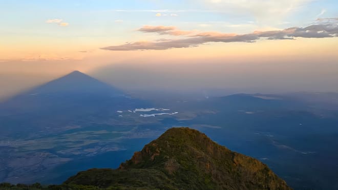 A high-altitude mountain peak overlooking a vast valley under a glowing orange and blue sunset sky.