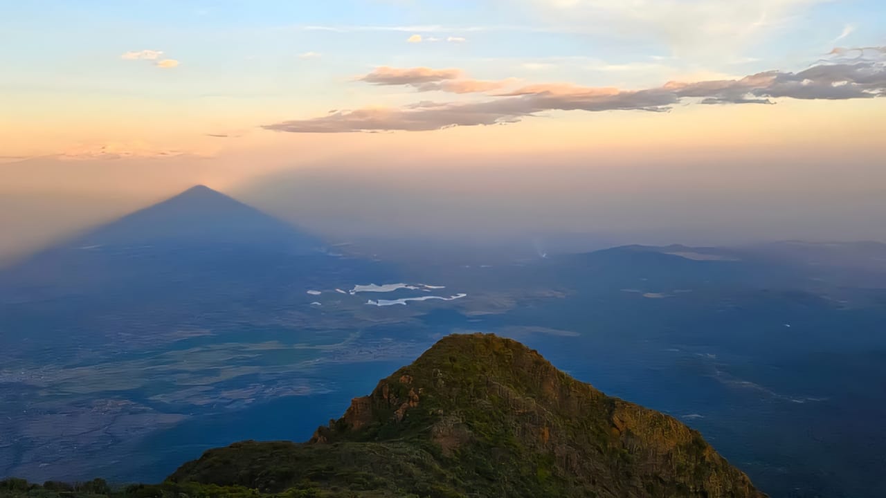 A high-altitude mountain peak overlooking a vast valley under a glowing orange and blue sunset sky.