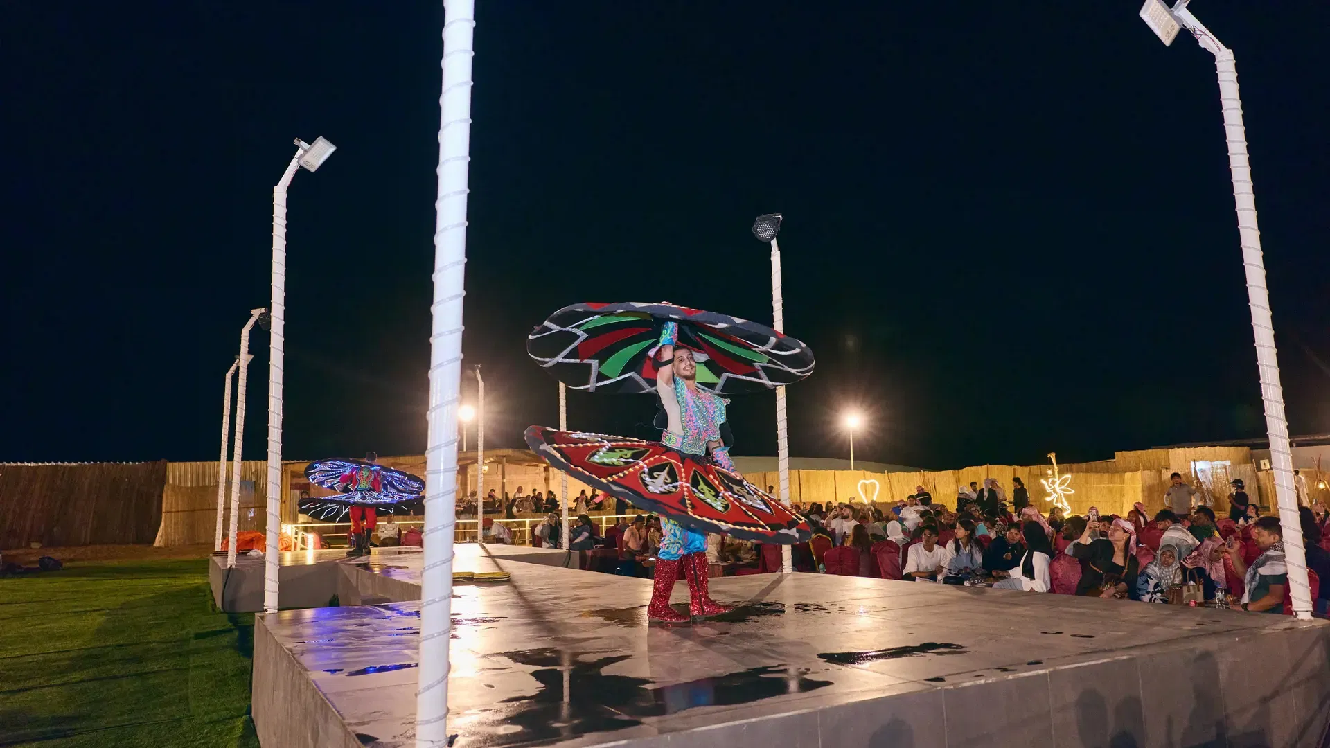 A performer spins a large, colorful circular skirt on stage under the night sky at Desert Safari + Al Faqaa Camp.