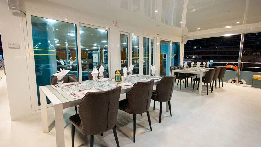 A light-colored dining table with grey chairs is set up near floor-to-ceiling glass doors, overlooking an exterior deck at night at Alexandra Sea Lounge.