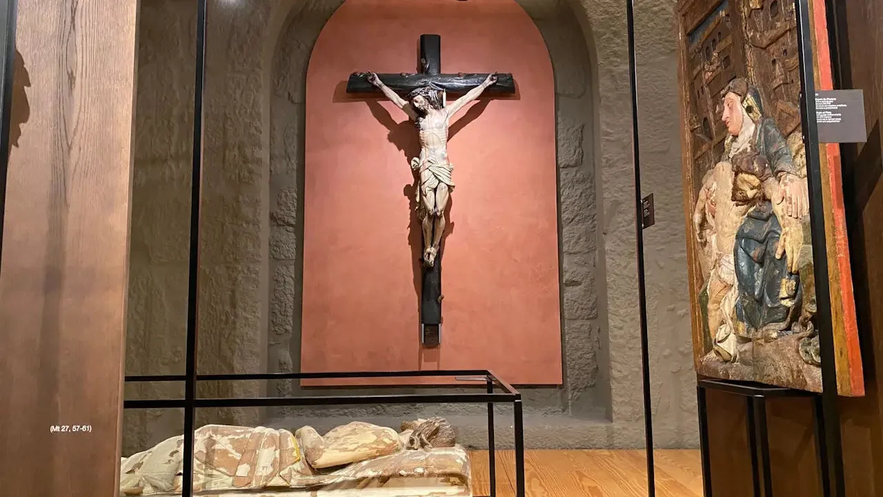 A large crucifix with a statue of Jesus is mounted on a pink arched wall above a glass case containing a recumbent figure at Clérigos Tower & Museum Ticket.