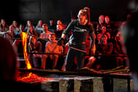 A female presenter in a "Lady Lava" shirt pours glowing orange molten lava with a metal rod in front of an engaged audience watching in awe.