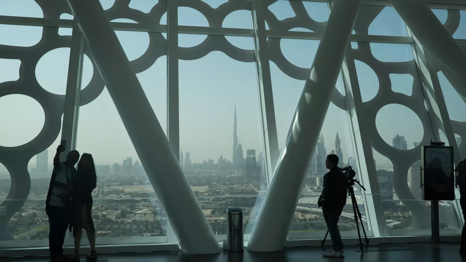 Visitors in silhouette taking a selfie and a photographer setting up a tripod against the patterned glass wall, the Burj Khalifa on the skyline beyond at Dubai Frame