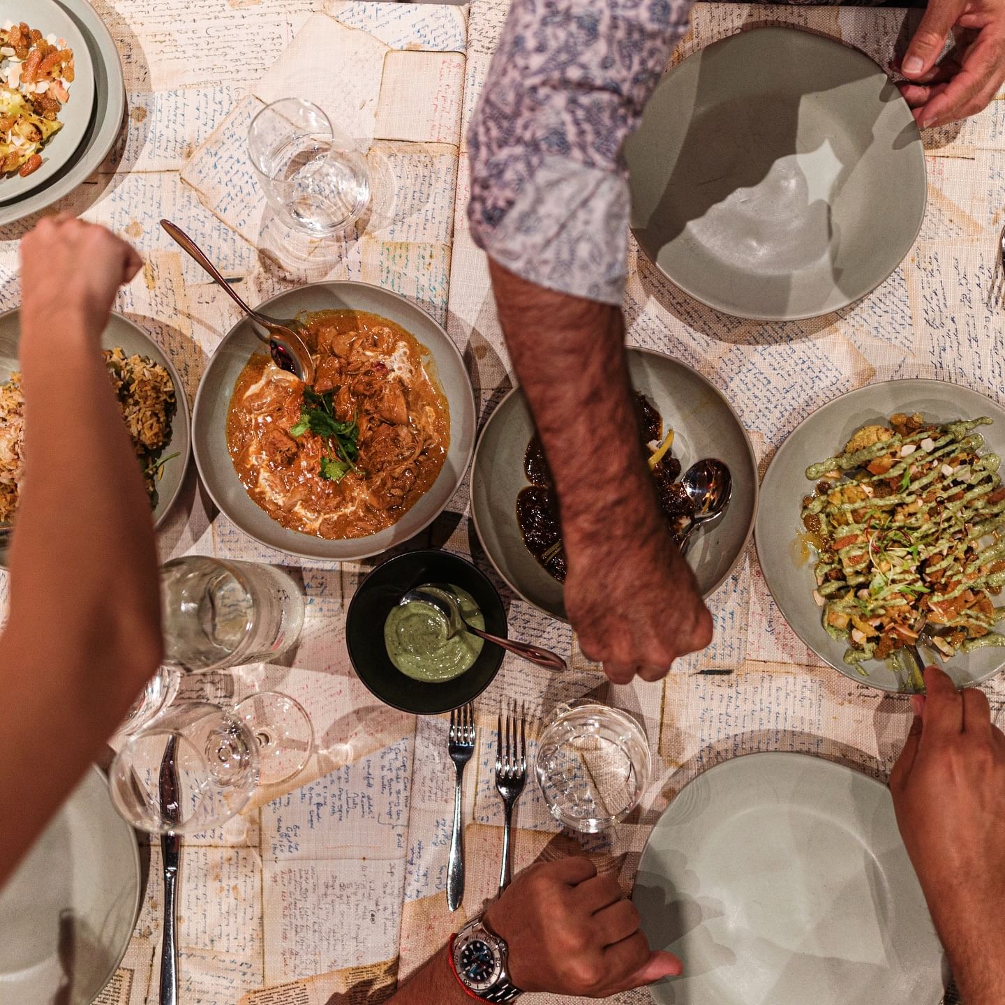 An overhead, close-up shot of a table set for a communal meal. Several hands are reaching for different plates of food.