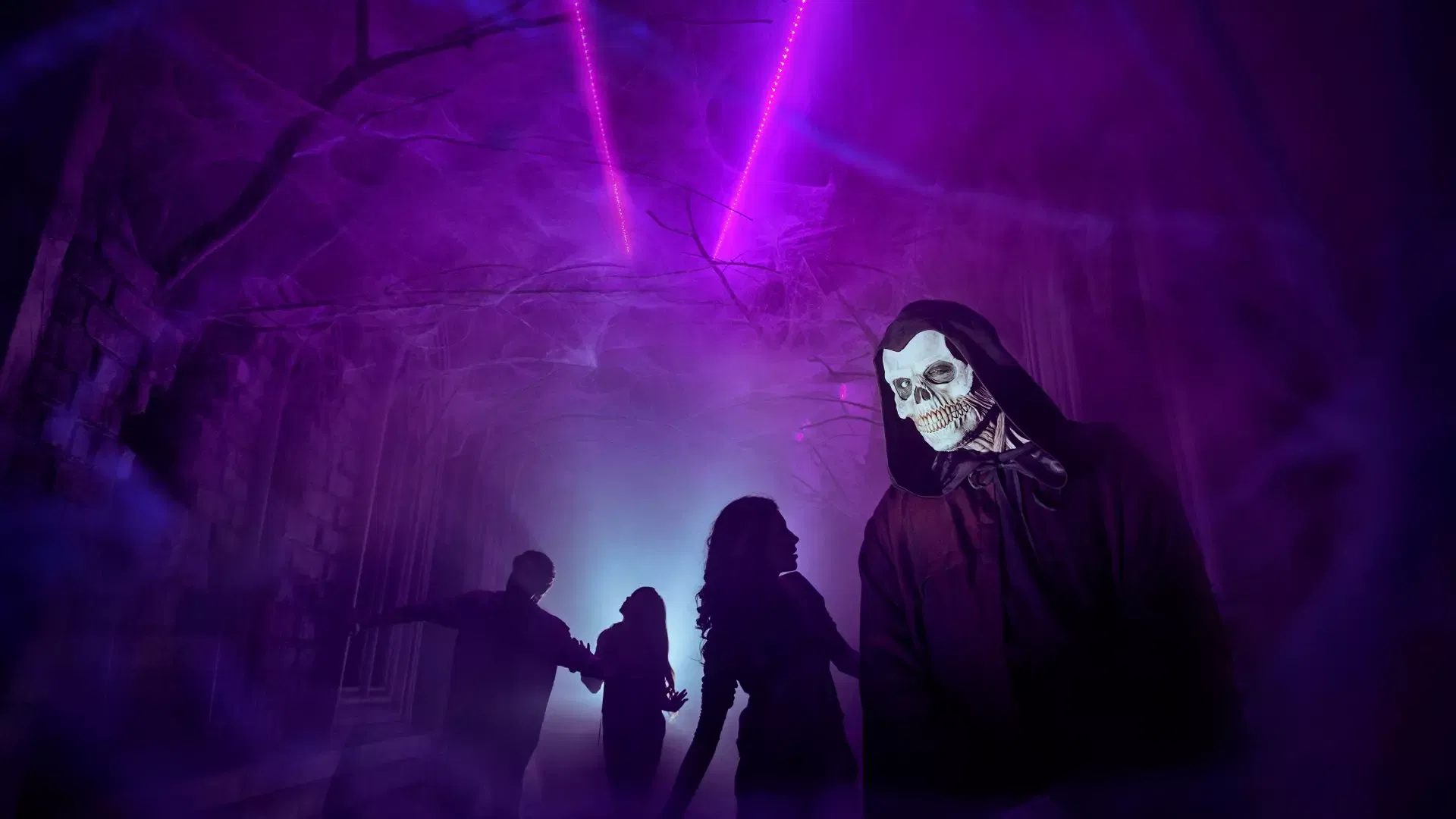 A person in a grim reaper mask stands in a purple-lit corridor with cobwebs as other visitors walk in the background at IMG Worlds of Adventure Theme Park.